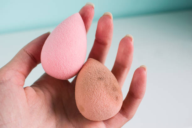 A hand holds a clean pink beauty blender next to a dirty orange one, highlighting cleaning differences.